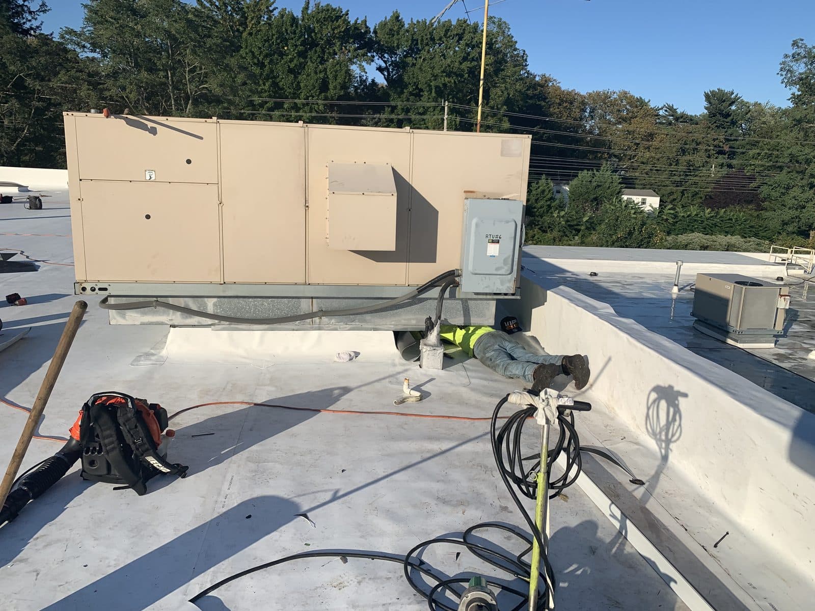 Rooftop HVAC unit on a flat commercial roof, white single-ply membrane flashed up its curb, a roofer working beneath it