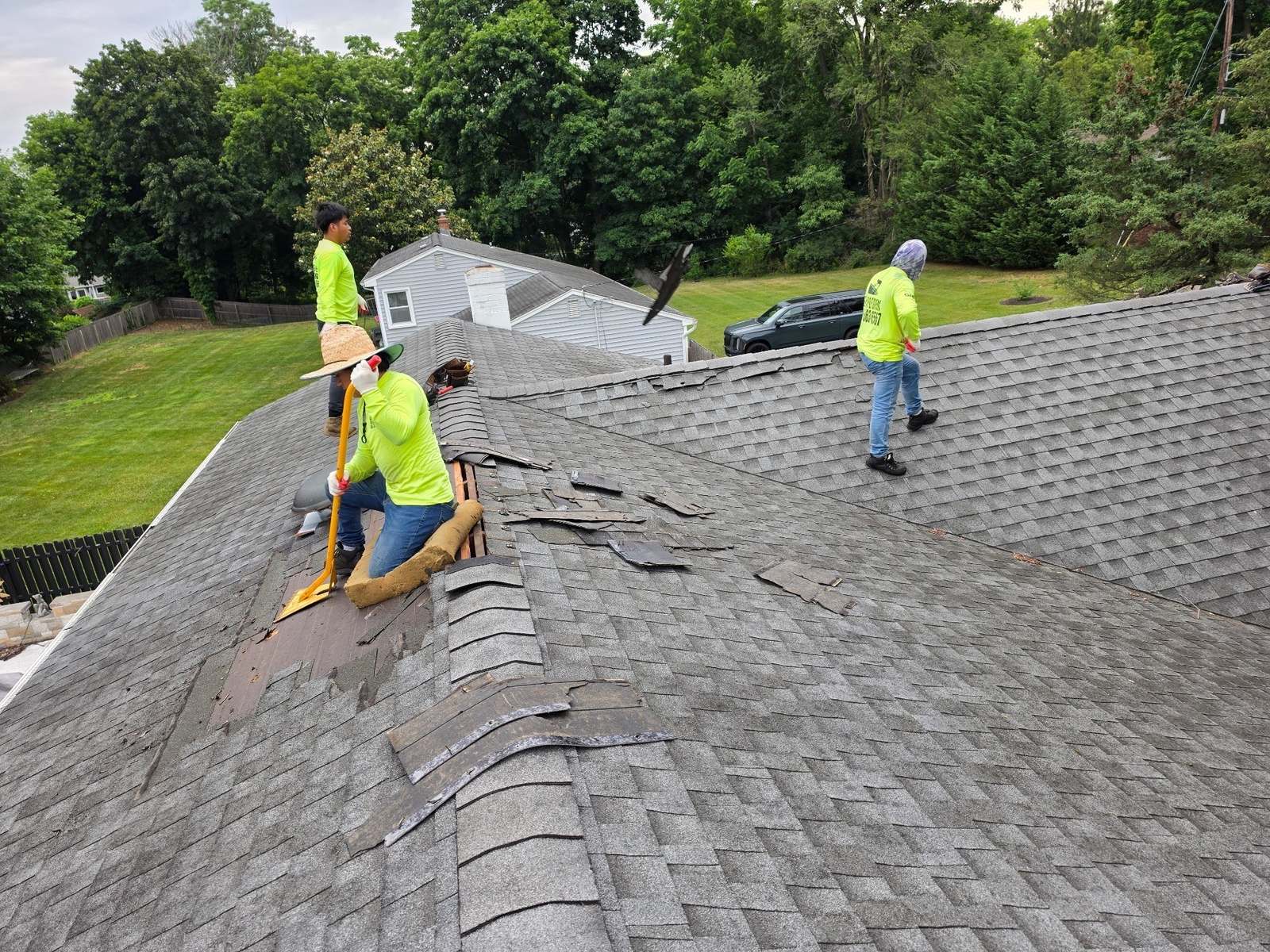 Roofers removing damaged gray architectural shingles along the hip of a pitched roof