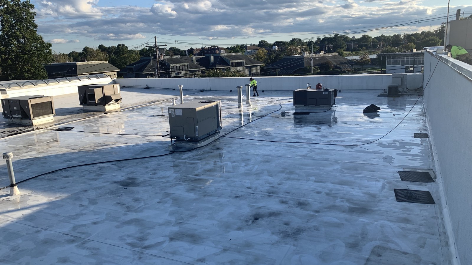 A large low-slope membrane roof over occupied space in New Jersey, carrying multiple rooftop HVAC units, drains and vents, with a Green Apple Roofing crew member working at the far parapet