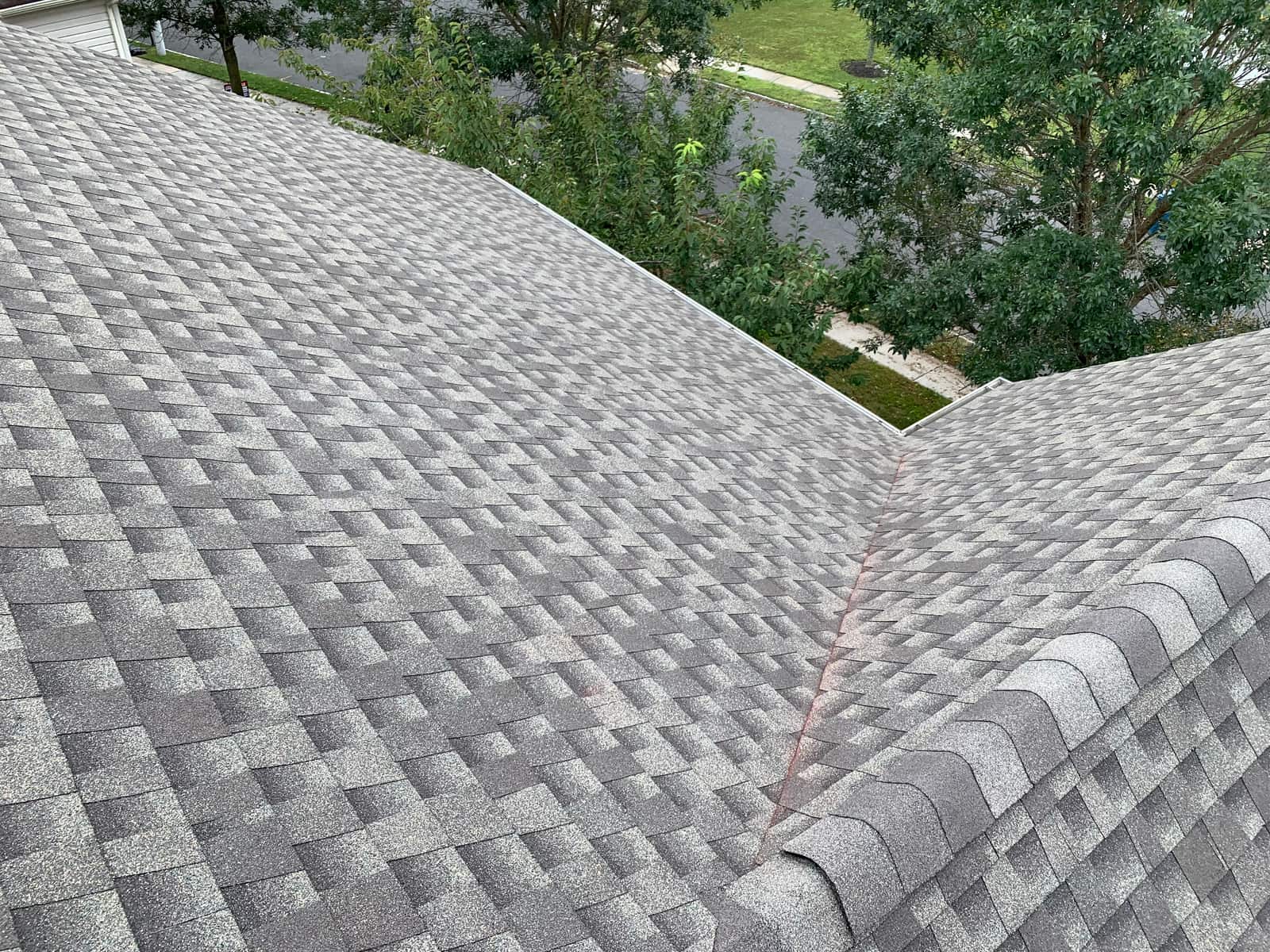 Finished gray architectural shingles where two roof slopes meet at a valley under a ridge cap, with trees and a lawn beyond
